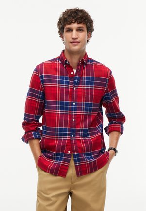 Junger Mann mit lockigem Haar, der ein rot-blau kariertes Hemd mit Knöpfen und beige Hosen trägt, steht mit den Händen in den Taschen vor weißem Hintergrund.