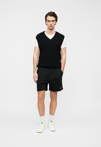 Μαύρο πλεκτό γιλέκο με λαιμόκοψη σε σχήμα V πάνω από λευκό T-shirt, συνδυασμένο με μαύρο σορτς και λευκά αθλητικά παπούτσια, στέκεται μπροστά σε απλό λευκό φόντο.