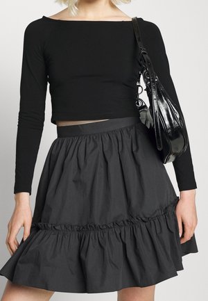 Kobieta ubrana w czarny crop top z długim rękawem i czarną spódnicę z wysokim stanem i falbankami, nosząca błyszczącą czarną torebkę na ramię.
