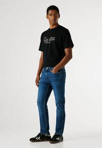 Černé bavlněné tričko s logem "Pepe Jeans", spárované s modrými džínami. Model má na sobě černé boty s bílými detaily. Jednoduchý design.