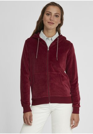 Burgundy zip-up hoodie in plush fabric with a drawstring hood, front pockets, and ribbed cuffs and hem. Worn over a striped shirt.