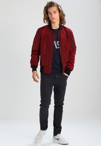 Chaqueta bomber burdeos con puños acanalados, bolsillo con cremallera en la manga, usada sobre una camiseta azul marino y jeans negros, combinada con zapatillas blancas.