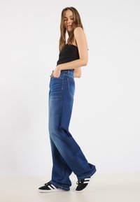Jean large bleu foncé en tissu denim, avec un design classique à cinq poches et des détails de couture minimalistes, associés à des baskets noires.