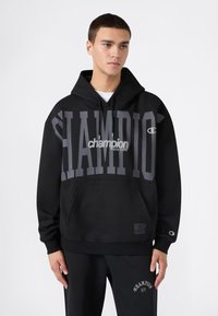 Sudadera negra con gran impresión en gris de "CHAMPION". Presenta un bolsillo tipo canguro, capucha ajustable con cordón y puños acanalados. Textura de tela suave.