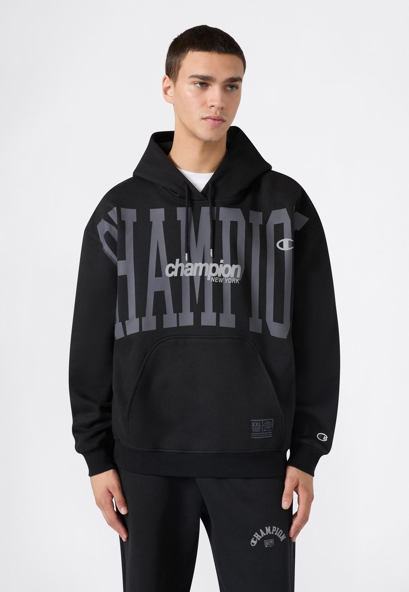 Sudadera negra con gran impresión en gris de "CHAMPION". Presenta un bolsillo tipo canguro, capucha ajustable con cordón y puños acanalados. Textura de tela suave.