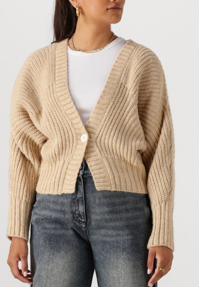 Vrouw in een beige chunky gebreide cropped cardigan over een witte top met hooggehakte vervaagde zwarte jeans, gouden sieraden zichtbaar om de nek en hand.