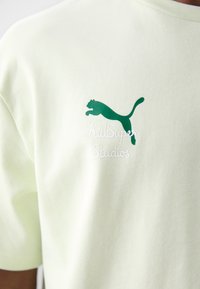 Camiseta de algodón verde claro con un logo de Puma verde oscuro y texto blanco "KidSuper Studios" bordado en el pecho.