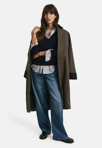 Over-sized olijfgroene trenchcoat over een marineblauwe gebreide trui, wit gestreept shirt en wijde blauwe jeans; zwarte schoenen met gestructureerde details.