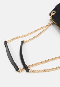 Musta nahkainen laukku, jossa on kultaketjuhihna. Suunnittelussa on kaksi kantohihnaa: yksi ketju ja yksi litteä nahka. Ylhäällä on vetoketjukiinnitys.