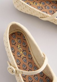 Scarpe piatte intrecciate color crema con una fascia e una fibbia. La soletta presenta uno sfondo marrone con motivi floreali blu.
