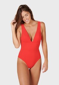 Maillot de bain une pièce rouge avec un décolleté en V profond, des bretelles larges et une texture lisse. Le design présente des découpes hautes sur les jambes et un style minimaliste.