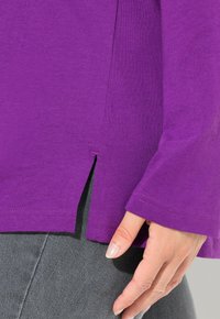 Top in cotone viola con maniche lunghe, con spacchi laterali all'orlo. Pantaloni grigi sono parzialmente visibili sotto la maglietta.