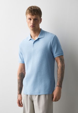 Ung mand med tatoveringer på begge underarme iført en lyseblå polo og beige bukser står op ad en hvid væg.