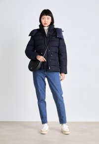 Námornícka pufovaná bunda s uzávermi na šnúrkach, vysokým golierom a detailmi na pleciach, kombinovaná s modrými džínami a bielymi teniskami. Čierna crossbody taška.