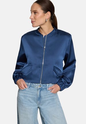 Femme aux cheveux foncés attachés en queue de cheval, portant une veste zippée bleue brillante et un jean bleu clair, regardant vers la gauche avec les mains dans les poches.