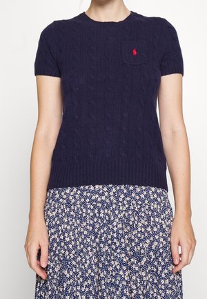 Femme portant un pull en maille torsadée bleu marine à manches courtes avec un petit logo rouge et une jupe à fleurs bleu marine avec des fleurs blanches et rose clair.