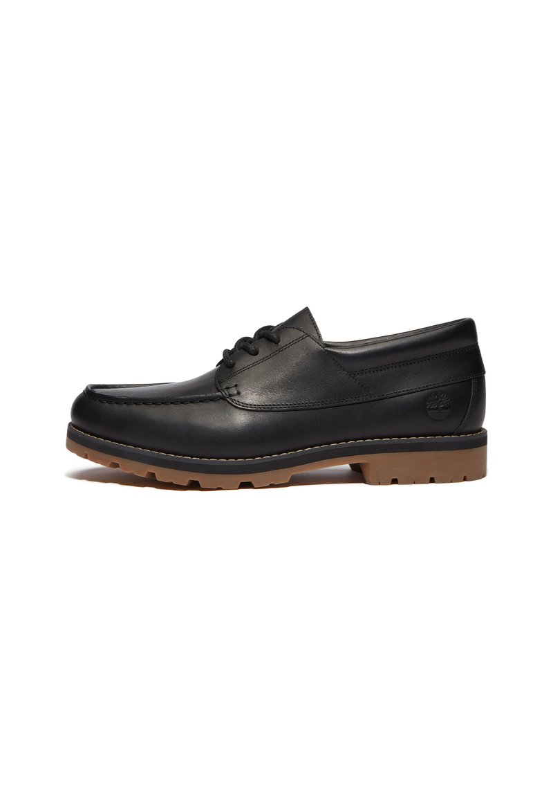 Zapato de cuero negro con punta redondeada, diseño con cordones, suela de goma y costuras en contraste. Presenta un logo en el talón.