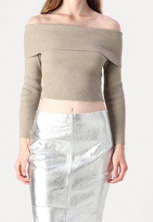 Vrouw draagt een beige off-shoulder geribbelde cropped trui en een hooggetailleerde metallic zilveren rok, staand met ontspannen armen.