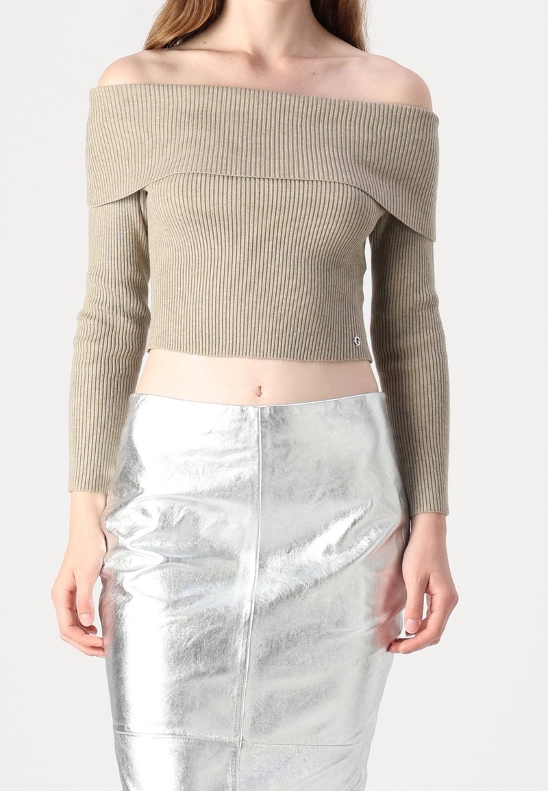 Femme portant un pull court côtelé beige à épaules dénudées et une jupe argentée métallisée taille haute, debout les bras détendus.