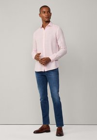 Camicia a maniche lunghe rosa chiaro con chiusura a bottoni, tessuto leggero e colletto a punta, abbinata a jeans di denim blu e scarpe marroni.
