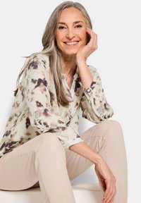Blusa leggera a maniche lunghe in beige chiaro con un motivo floreale astratto marrone; bottoni ai polsini; modello seduto che sorride.