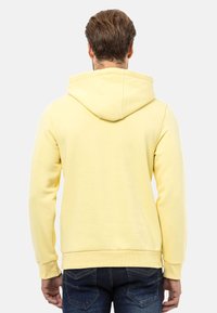 Gelbe Kapuzenjacke mit Kordelzug und gerippten Bündchen. Weicher Stoff mit lockerer Passform, kombiniert mit dunkelblauen Jeans.