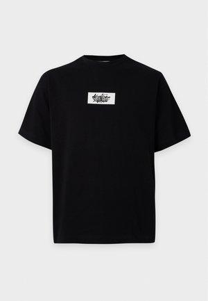 Jeune homme debout, les mains dans le dos, portant un t-shirt noir à motif graphique, un pantalon large et sombre, et des baskets claires sur un fond uni.