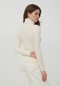 Maglione a collo alto a costine in crema chiaro, con maniche lunghe e design aderente. Texture liscia con dettagli di cuciture sottili.