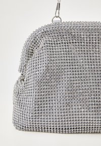 Sac à main argenté orné d'une garniture de cristal circulaire, présentant une forme structurée et une fermeture à glissière sur le dessus. Doublure en tissu lisse visible.