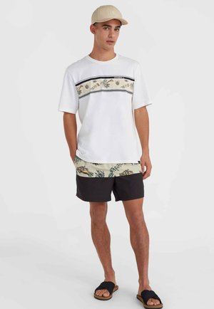 T-shirt graphique blanche avec imprimé tropical et accents noirs, associée à un short noir avec un motif de plage et des sandales.