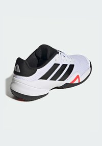 Bela športna obutev s črnimi poudarki in tremi črnimi črtami. Strukturiran zgornji del, oblazinjen podplat, rdeč detajl na strani in črn gumijast podplat.