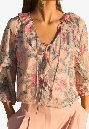 Vrouw die een doorschijnende blouse met bloemenprint draagt, met ruches en een striksluiting bij de hals, gecombineerd met een high-waist roze broek en grote groene oorbellen.