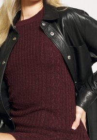 Musta nahkatakki metallinapeilla burgundinvärisen kaapelikudottu neuleen päällä. Neuleessa on raidoitettu pinta, mikä korostaa sen istuvaa muotoilua.