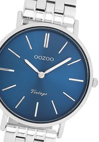 Cadran de montre bleu rond avec des index horaires argentés, doté d'un bracelet en métal argenté et du texte "OOZOO Vintage". Design lisse et minimaliste.