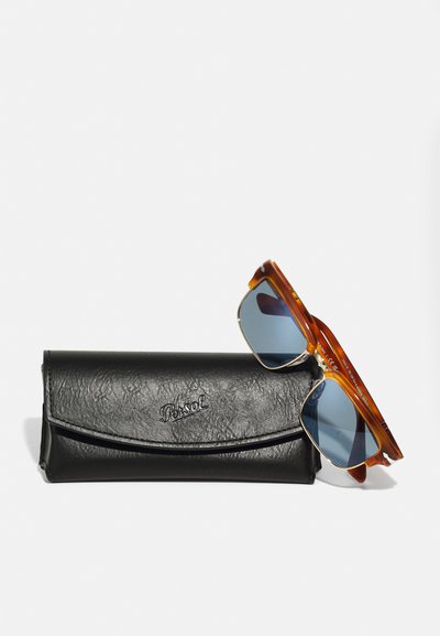 Óculos de sol de tartaruga com lentes azuis e detalhes em dourado, repousados sobre uma caixa de couro preto texturizado com logotipo em relevo.