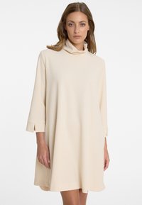 Beige, knielanges Kleid aus weichem Stoff mit hohem Kragen und Dreiviertelärmeln, die überlappende Manschetten und eine lockere Passform aufweisen.