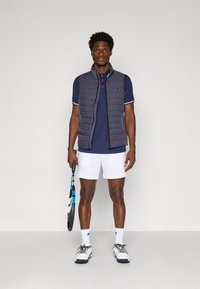 Marinblå ärmlös väst med quiltat mönster över marinblå piké, vita shorts och sportskor. Håller en tennisracket, accessoriserad med vita strumpor.