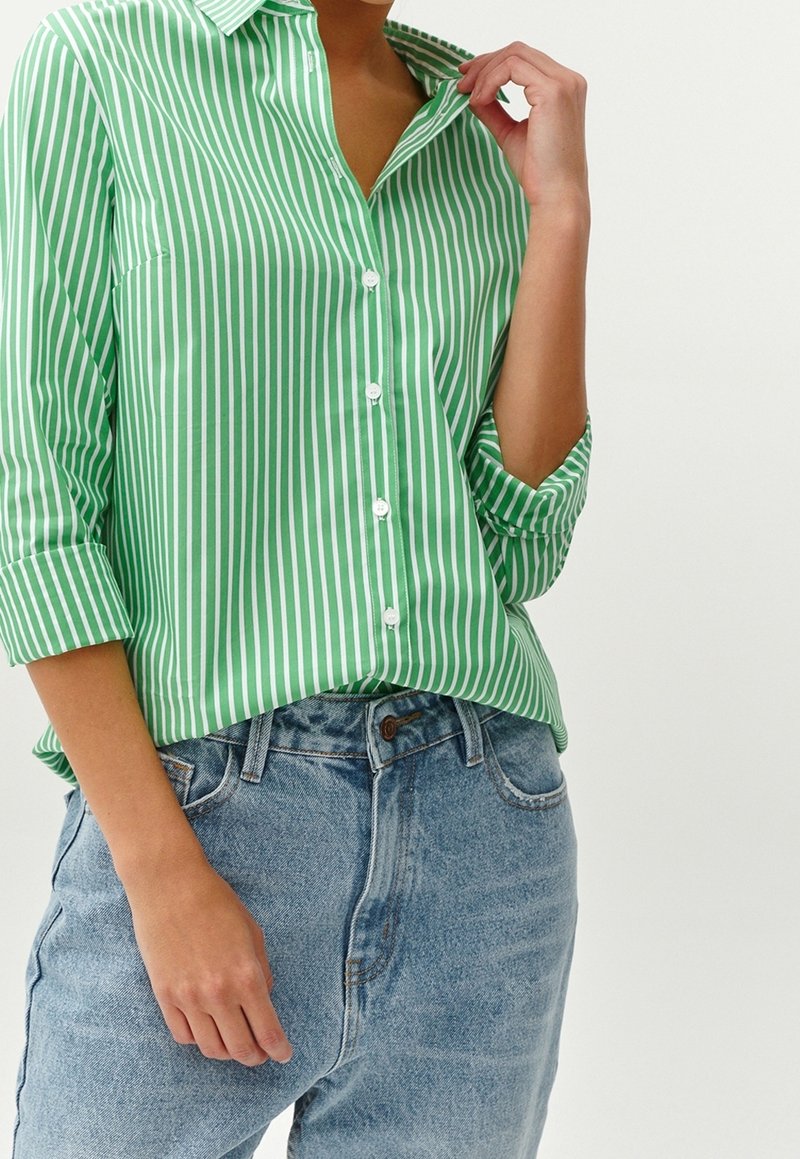 Personne portant une chemise à rayures verticales vertes et blanches à boutons avec les manches retroussées et un jean denim bleu clair à taille haute.