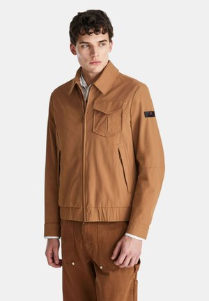 Giovane uomo con capelli corti e ricci che indossa una giacca marrone con zip e tasca frontale con patta e pantaloni marroni abbinati, guarda di lato.