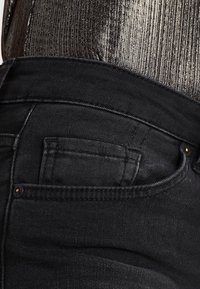 Vaqueros de mezclilla negra con una cinturilla alta, que cuentan con un pequeño bolsillo trasero y detalles de costura sutiles. La tela tiene una textura suave.