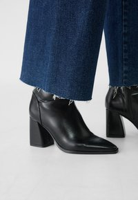 Black leather high-heeled ankle boots with a pointed toe, square block heel, and subtle stitching detail, paired with frayed hem dark denim pants.