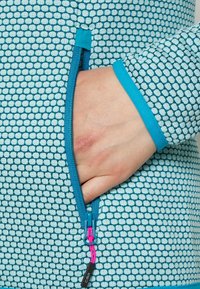 Giacca azzurra chiusa con zip che presenta un motivo testurizzato, bordi turchesi a contrasto e un tirazip rosa. Una tasca è visibile sul lato.
