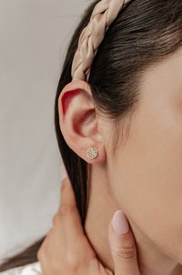 Orecchini a bottone in oro con un motivo geometrico, indossati all'orecchio di un modello, abbinati a una fascia intrecciata per capelli e smalto per unghie in tonalità neutre.