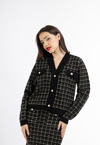 Cardigan nero e crema con texture, scollatura a V, due tasche con patta e bottoni perlati sul davanti e sui polsini. Dettagli del tessuto lavorato a maglia visibili.