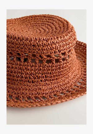 Sombrero de paja trenzada en color terracota con una corona redondeada y un ala ancha y flexible. Presenta patrones de tejido abierto alrededor de los lados.