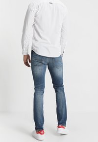 Bílá košile s dlouhým rukávem, modré džíny a bílé tenisky s červenými akcenty; uvolněný střih s detaily na zadních kapsách.