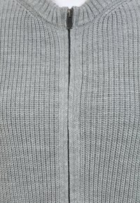 Cárdigan de punto gris con cierre de cremallera, que presenta una textura acanalada y un cuello redondo. Tejido suave con un patrón uniforme.