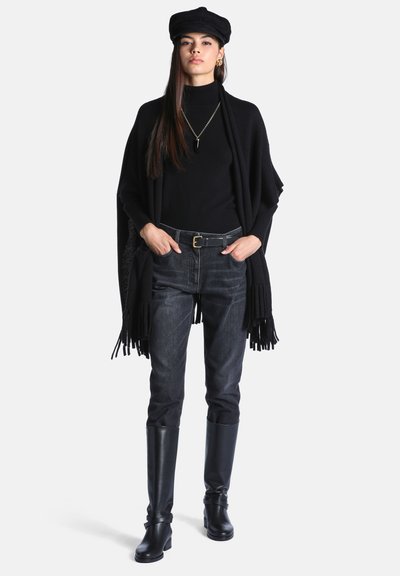 Jeune femme portant un col roulé noir, un châle à franges, une ceinture noire, un jean foncé, des bottes noires montantes jusqu'aux genoux, une casquette noire et des bijoux en or, debout avec les mains dans les poches.