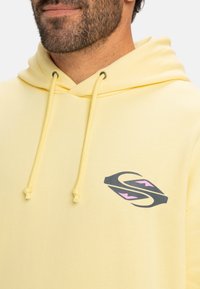 Yellow hoodie with a drawstring, featuring a graphic logo in black and pink. The fabric appears soft with a smooth texture.