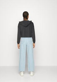 Klippt svart huvtröja med snörning i huvan, matchad med ljusblå vida jeans med cargo-fickor. Modell vänder ryggen mot kameran.